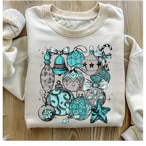 Peut inclure: Un sweat-shirt cr&egrave;me avec un imprim&eacute; graphique d'ornements de No&euml;l bleu sarcelle et argent. Les ornements sont dessin&eacute;s dans un style vintage et comprennent des cloches, des &eacute;toiles et des cannes en sucre.
