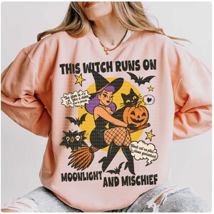 May include: A pale pink sweatshirt with a Halloween design. The graphic includes a witch on a broom, a black cat, and a pumpkin. The text on the sweatshirt says "THIS WITCH RUNS ON MOONLIGHT AND MISCHIEF."