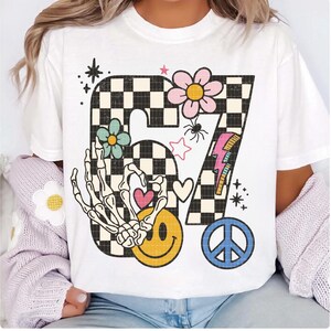 May include: White t-shirt featuring a large, retro-style graphic. The design includes the number "67" in a black and white checkerboard pattern, adorned with flowers, a skeleton hand, a smiley face, a peace sign, and a lightning bolt.