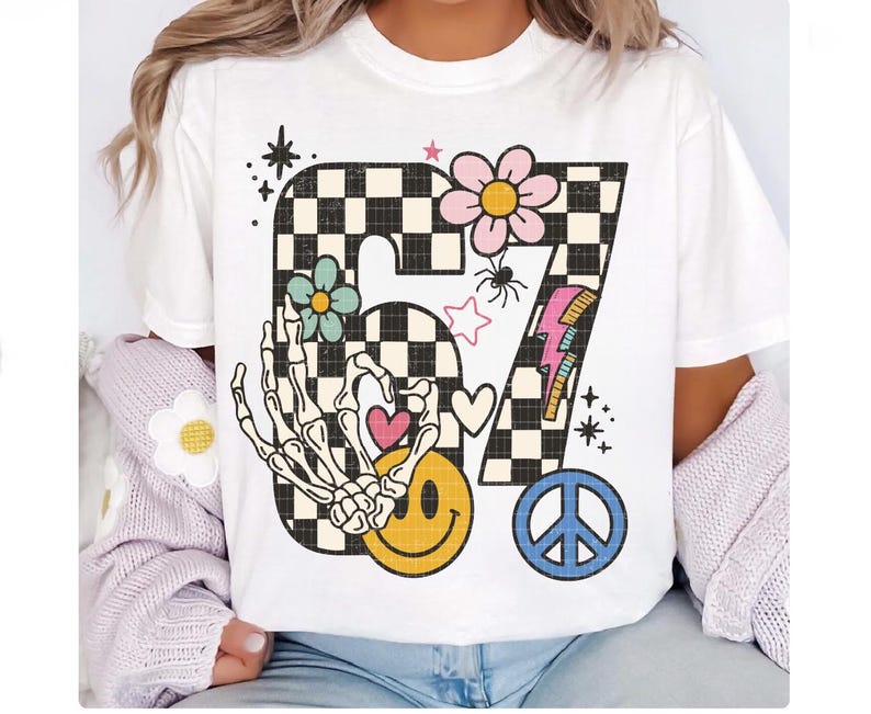 May include: White t-shirt featuring a large, retro-style graphic. The design includes the number "67" in a black and white checkerboard pattern, adorned with flowers, a skeleton hand, a smiley face, a peace sign, and a lightning bolt.