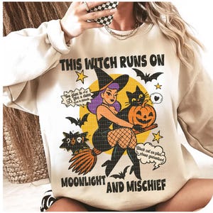 May include: Cream sweatshirt with a Halloween graphic. The design shows a cartoon witch with purple hair, a black cat, and a jack-o'-lantern. Text reads "THIS WITCH RUNS ON MOONLIGHT AND MISCHIEF". Bats and stars complete the design.