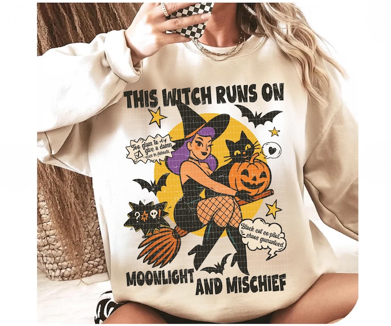 May include: Cream sweatshirt with a Halloween graphic. The design shows a cartoon witch with purple hair, a black cat, and a jack-o'-lantern. Text reads "THIS WITCH RUNS ON MOONLIGHT AND MISCHIEF". Bats and stars complete the design.