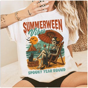 May include: White t-shirt with a colorful graphic featuring a skeleton relaxing on a beach under an umbrella, holding a glass of wine. The text reads "SUMMERWEEN Vibes" and "SPOOKY YEAR ROUND". A black cat sits nearby.