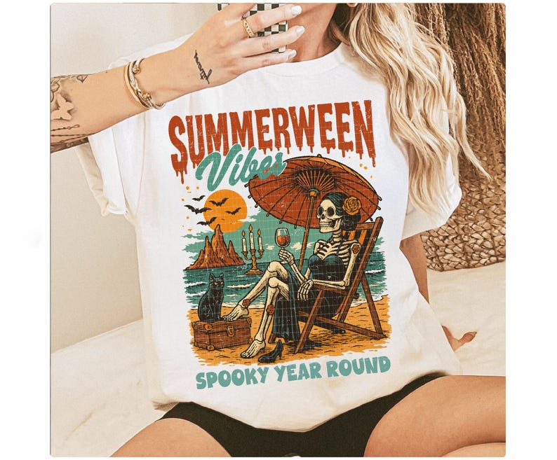 May include: White t-shirt with a colorful graphic featuring a skeleton relaxing on a beach under an umbrella, holding a glass of wine. The text reads "SUMMERWEEN Vibes" and "SPOOKY YEAR ROUND". A black cat sits nearby.