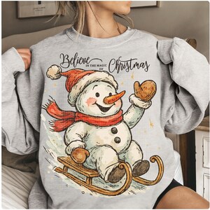 Op de afbeelding: Een lichtgrijze sweatshirt met een vrolijk sneeuwpopontwerp. De sneeuwpop draagt een rode sjaal, hoed en wanten en rijdt op een houten slee. De tekst "Believe in the Magic of Christmas" staat boven de sneeuwpop.