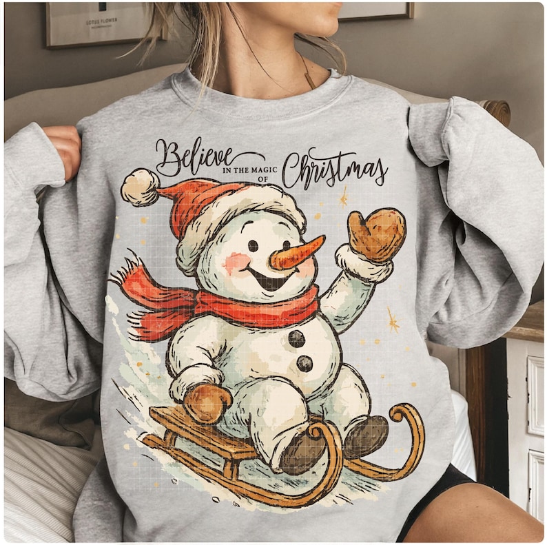Op de afbeelding: Een lichtgrijze sweatshirt met een vrolijk sneeuwpopontwerp. De sneeuwpop draagt een rode sjaal, hoed en wanten en rijdt op een houten slee. De tekst "Believe in the Magic of Christmas" staat boven de sneeuwpop.