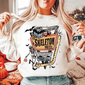 Puede incluir: Sudadera blanca con un letrero de estilo retro de "The Skeleton Motel". El dise&ntilde;o incluye un esqueleto, murci&eacute;lagos y el texto "Old Graveyard Road" y "No Escape Since 1690". Un gr&aacute;fico tem&aacute;tico de Halloween.