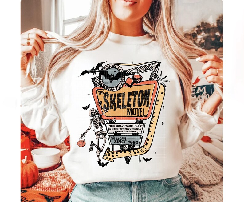 Puede incluir: Sudadera blanca con un letrero de estilo retro de "The Skeleton Motel". El dise&ntilde;o incluye un esqueleto, murci&eacute;lagos y el texto "Old Graveyard Road" y "No Escape Since 1690". Un gr&aacute;fico tem&aacute;tico de Halloween.