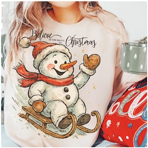 Op de afbeelding: Een lichtroze sweatshirt met een vrolijke sneeuwman illustratie. De sneeuwman draagt een rode hoed en sjaal en zit op een houten slee. De tekst "Believe in the magic of Christmas" staat boven de sneeuwman. Een mok met een boomontwerp is zichtbaar.