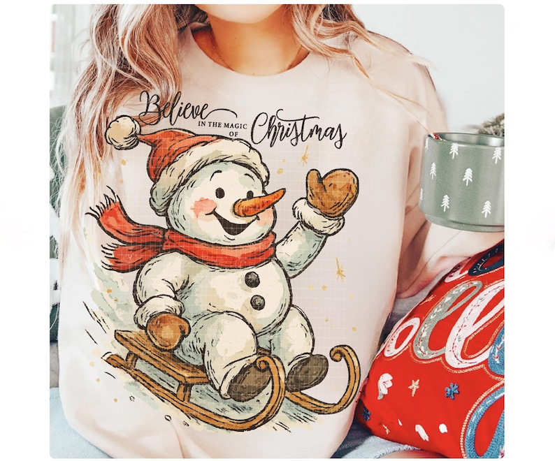 Op de afbeelding: Een lichtroze sweatshirt met een vrolijke sneeuwman illustratie. De sneeuwman draagt een rode hoed en sjaal en zit op een houten slee. De tekst "Believe in the magic of Christmas" staat boven de sneeuwman. Een mok met een boomontwerp is zichtbaar.
