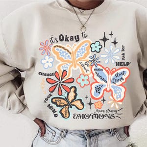 May include: A beige sweatshirt with a colorful graphic print of butterflies and flowers. The text on the sweatshirt reads "it's Okay to... CHANGE YOUR MIND... Be different... make Mistakes... ask for 'HELP'... Start Over... have strong EMOTIONS".