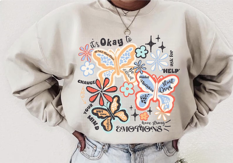 May include: A beige sweatshirt with a colorful graphic print of butterflies and flowers. The text on the sweatshirt reads "it's Okay to... CHANGE YOUR MIND... Be different... make Mistakes... ask for 'HELP'... Start Over... have strong EMOTIONS".
