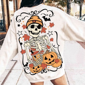May include: White sweatshirt with a skeleton wearing a beanie holding a pumpkin and a cup of coffee. The design also includes fall leaves and stars.