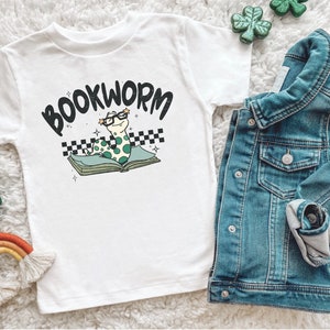 Pu&ograve; includere: Maglietta bianca con un verme verde che indossa gli occhiali e legge un libro. La maglietta dice "BOOKWORM" in lettere nere.