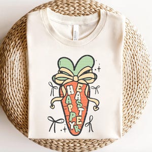 May include: A beige t-shirt with a graphic of a carrot with a bow on top. The carrot has the words "Happy Easter" written on it.