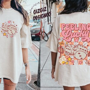 May include: Oversized beige t-shirt with a retro graphic of dice, stars, and the words "Feeling Lucky." The front features dice and stars. The back has the phrase "Feeling Lucky" in pink and orange, with dice and cactus illustrations.