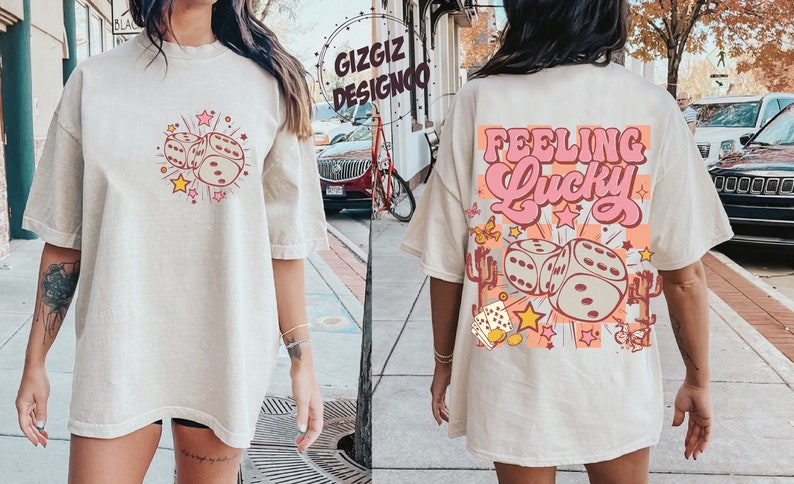 May include: Oversized beige t-shirt with a retro graphic of dice, stars, and the words "Feeling Lucky." The front features dice and stars. The back has the phrase "Feeling Lucky" in pink and orange, with dice and cactus illustrations.