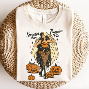Peut inclure: T-shirt cr&egrave;me avec une illustration r&eacute;tro d'une femme en costume d'Halloween, entour&eacute;e de citrouilles sculpt&eacute;es. Le texte "Sweeter than Pumpkin Pie" est au-dessus de l'illustration. Le t-shirt est en mati&egrave;re douce.