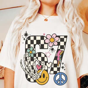 May include: Cream-colored t-shirt featuring a large, retro-style graphic. The graphic includes a checkered number '67' with a flower, spider, lightning bolt, and peace sign. A skeleton hand forms a heart with a smiley face.