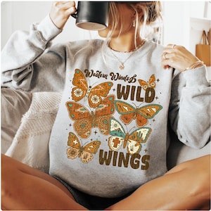 以下が含まれることがあります： 「Western Winds & Wild Wings」の文字と蝶のデザインが施されたグレーのスウェットシャツ。蝶はオレンジ、茶色、ティール色で描かれています。スウェットシャツはクルーネックです。