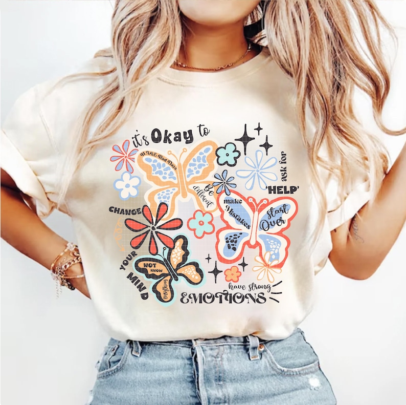 May include: A white t-shirt with a colorful graphic design featuring butterflies and flowers. The text on the shirt reads "it's Okay to" and includes phrases like "Change your mind", "Be different", "Make mistakes", "Start over", "Have strong emotions", and "Ask for help".