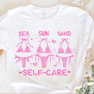 May include: White t-shirt with a pink graphic of three bikini tops and bottoms. The words "SEA SUN SAND" are above the bikinis, and "SELF-CARE" is below. The bikini design has a zebra stripe pattern.