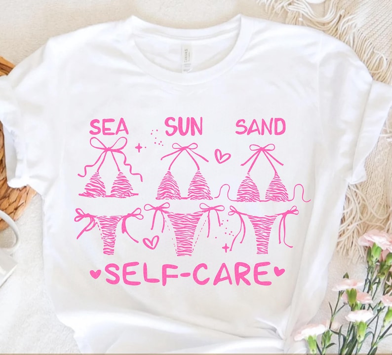 May include: White t-shirt with a pink graphic of three bikini tops and bottoms. The words "SEA SUN SAND" are above the bikinis, and "SELF-CARE" is below. The bikini design has a zebra stripe pattern.