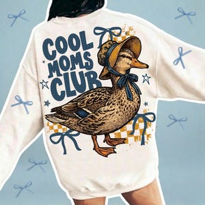 May include: Cream-colored sweatshirt with a graphic of a duck wearing a bonnet and the text "COOL MOMS CLUB" in navy blue. The duck is brown and tan with blue accents. Blue bows and stars are scattered around the design.