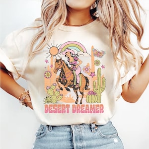Pu&ograve; includere: Una maglietta bianca con un design grafico retr&ograve; che presenta una donna a cavallo in un paesaggio desertico. Il grafico include un sole, un arcobaleno, un cactus, fiori e farfalle. Il testo "DESERT DREAMER" &egrave; stampato sotto il grafico.
