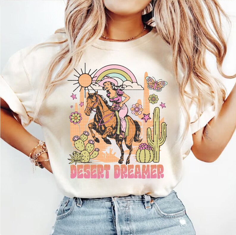 Pu&ograve; includere: Una maglietta bianca con un design grafico retr&ograve; che presenta una donna a cavallo in un paesaggio desertico. Il grafico include un sole, un arcobaleno, un cactus, fiori e farfalle. Il testo "DESERT DREAMER" &egrave; stampato sotto il grafico.