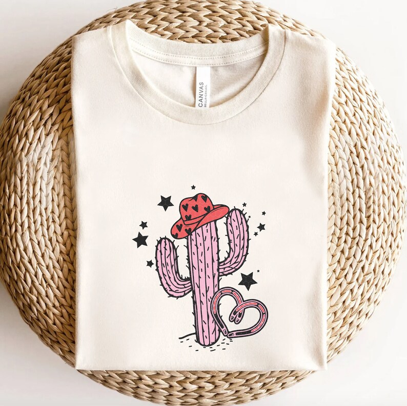 May include: A beige t-shirt with a pink cactus wearing a red cowboy hat with black hearts. The cactus is surrounded by black stars and a pink horseshoe with black hearts.