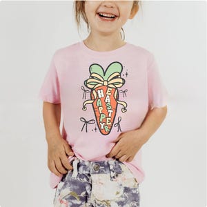 May include: Pink short-sleeved t-shirt with a cartoon carrot graphic. The carrot has a green bow and the words "Happy Easter" written on it.