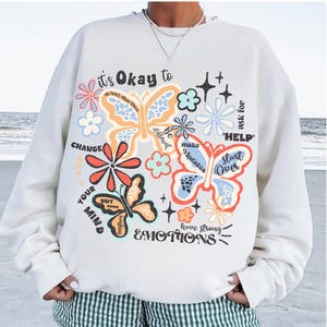 May include: White sweatshirt with a colorful butterfly graphic and the text "it's Okay to" and "ask for HELP". The graphic also includes flowers and the text "CHANGE YOUR MIND", "Be different", "make Mistakes", "Start Over", and "have strong EMOTIONS".