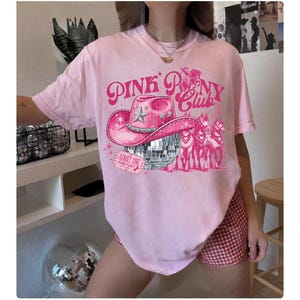 May include: Pink t-shirt with a graphic design featuring a pink cowboy hat, a disco ball, and the text "Pink Pony Club." The design also includes a ticket that says "Admit One." The shirt is a light pink color.