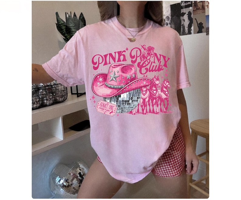 May include: Pink t-shirt with a graphic design featuring a pink cowboy hat, a disco ball, and the text "Pink Pony Club." The design also includes a ticket that says "Admit One." The shirt is a light pink color.