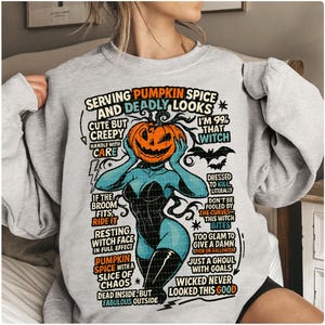May include: A light gray sweatshirt with a Halloween-themed graphic. The graphic features a woman with a pumpkin head, surrounded by text that reads "Serving Pumpkin Spice and Deadly Looks." The design includes bats and witch-related phrases.