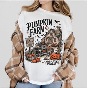 Peut inclure: Un t-shirt blanc avec une impression graphique repr&eacute;sentant une ferme de citrouilles, une maison, un camion et des citrouilles. Le texte indique PUMPKIN FARM et MAGICALLY GROWN, avec des panneaux PICK YOUR PUMPKIN FRESHLY GROWN. Le motif inclut des chauves-souris.