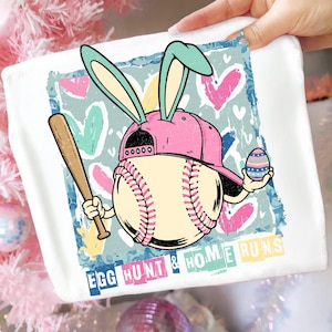 May include: White graphic tee featuring a baseball with bunny ears and a pink cap, holding a bat and an Easter egg. The design includes the text "EGG HUNT & HOME RUNS" with colorful hearts in the background.