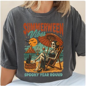 May include: Dark gray t-shirt with a graphic featuring a skeleton relaxing on a beach chair under an orange umbrella. The design includes the text "SUMMERWEEN Vibes" and "SPOOKY YEAR ROUND". A black cat sits nearby.