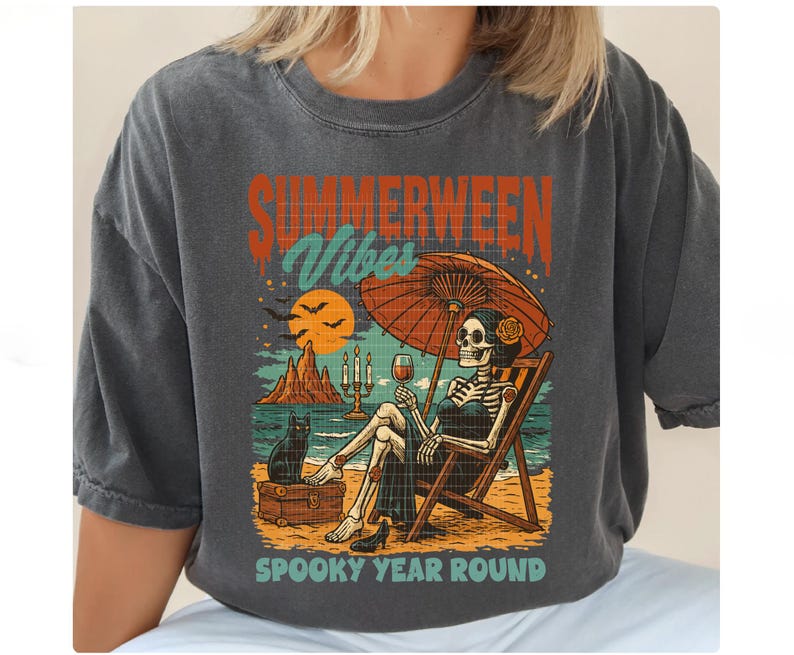 May include: Dark gray t-shirt with a graphic featuring a skeleton relaxing on a beach chair under an orange umbrella. The design includes the text "SUMMERWEEN Vibes" and "SPOOKY YEAR ROUND". A black cat sits nearby.