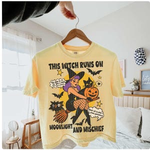 May include: A pale yellow t-shirt with a Halloween graphic. The design includes a witch on a broom, a black cat, bats, and a pumpkin. Text on the shirt reads "THIS WITCH RUNS ON MOONLIGHT AND MISCHIEF".