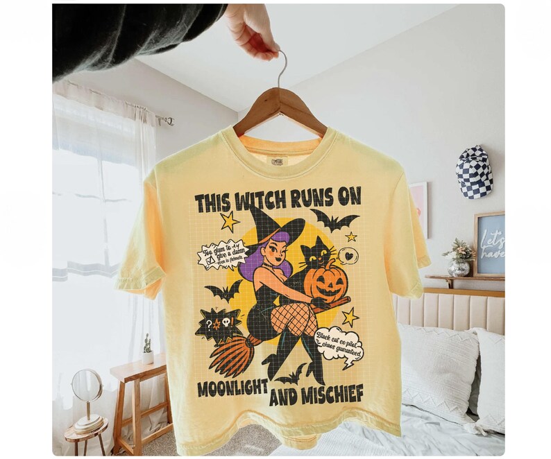 May include: A pale yellow t-shirt with a Halloween graphic. The design includes a witch on a broom, a black cat, bats, and a pumpkin. Text on the shirt reads "THIS WITCH RUNS ON MOONLIGHT AND MISCHIEF".
