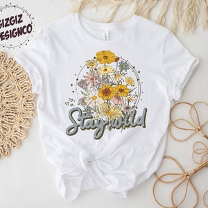 May include: White t-shirt with a floral design and the text "Stay Wild" in blue lettering.