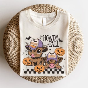 Puede incluir: Una camiseta beige con un gr&aacute;fico de dos vacas de dibujos animados que llevan sombreros de vaquero y sostienen calabazas. El texto "Howdy Fall" est&aacute; por encima de las vacas.