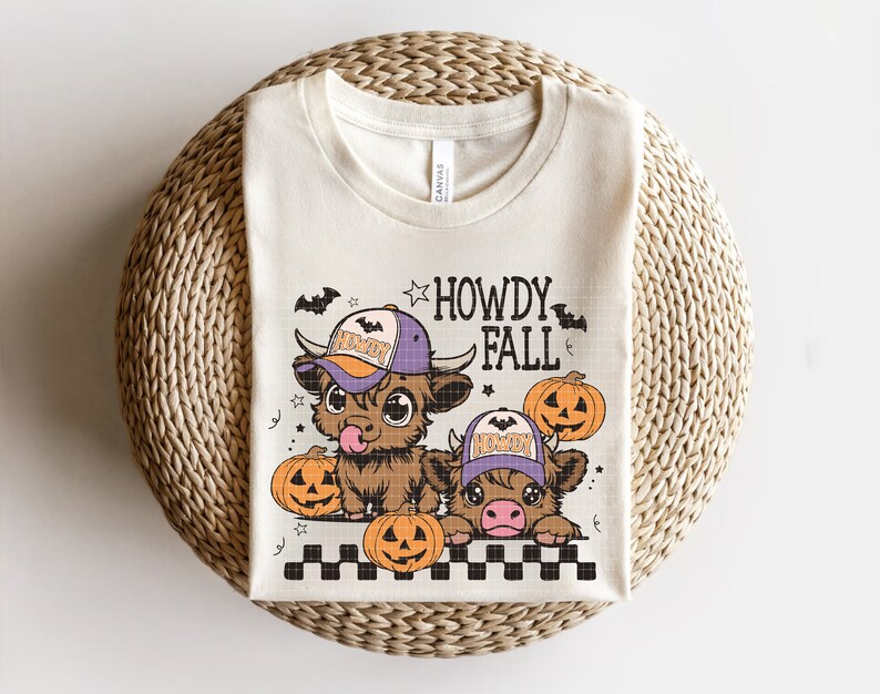 Puede incluir: Una camiseta beige con un gr&aacute;fico de dos vacas de dibujos animados que llevan sombreros de vaquero y sostienen calabazas. El texto "Howdy Fall" est&aacute; por encima de las vacas.