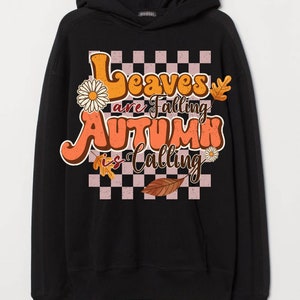 Op de afbeelding: Zwart hoodie met een grafische print van een geruit patroon, bladeren en de tekst "Leaves are falling, Autumn is calling".
