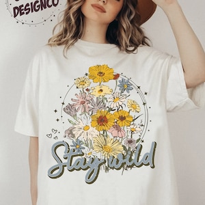 May include: White t-shirt with a floral design and the text "Stay Wild" in a green and blue font.