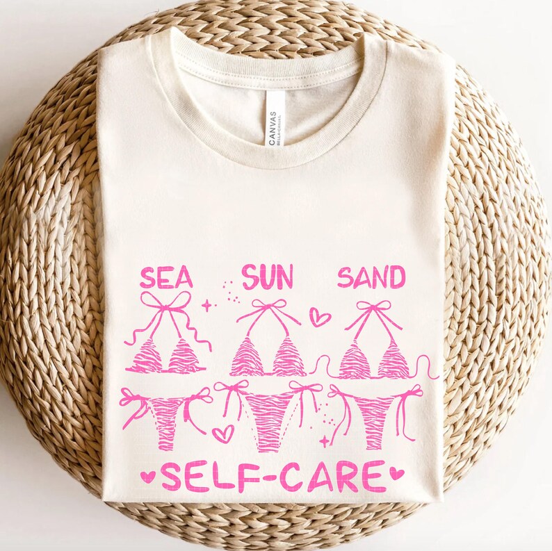 May include: Cream-colored t-shirt with a pink graphic of three bikini tops and bottoms. The words "SEA SUN SAND SELF-CARE" are printed in pink. The shirt is made of a soft material and is perfect for summer.