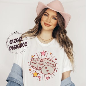 May include: White t-shirt featuring a graphic of two dice with red details, surrounded by stars and lines. The text "GIZGIZ DESIGNCO" is in a circle on the left side of the image. The shirt is worn by a person wearing a pink hat.