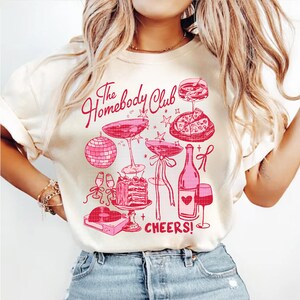 May include: A white t-shirt with a pink graphic design that says "The Homebody Club" and features a disco ball, champagne glasses, a bottle of wine, a slice of pizza, a cake, and a record player. The text "Cheers!" is also included in the design.